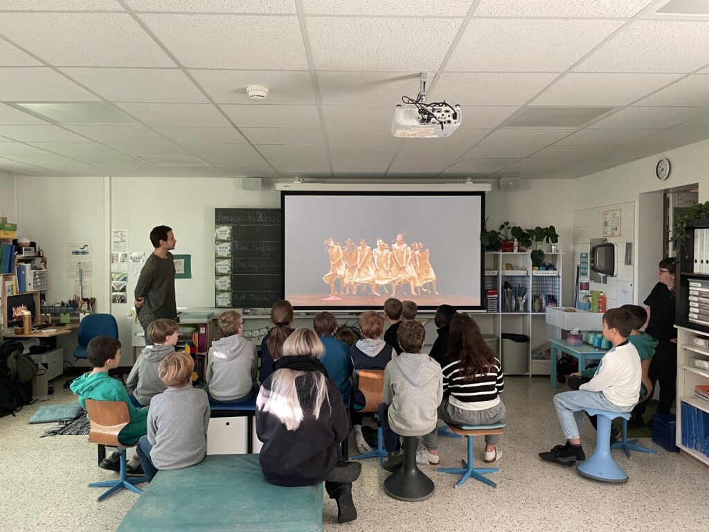 Une classe d'enfants regardent un écran sur lequel se trouvent des danseurs. 